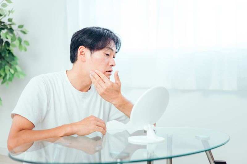 Tゾーンはベタつくのに頬は粉を吹くような乾燥を感じ、肌の違和感を確認する男性。水分保持力の低下により、夕方になると肌がゴワつくインナードライ特有の症状を示すシーン。