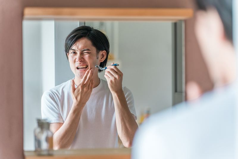 髭剃り直後の肌の赤みやヒリつきに悩み、顔を歪ませる男性。角質層が削られ、外部刺激に対して一時的に非常にデリケートになっている肌状態を表現。