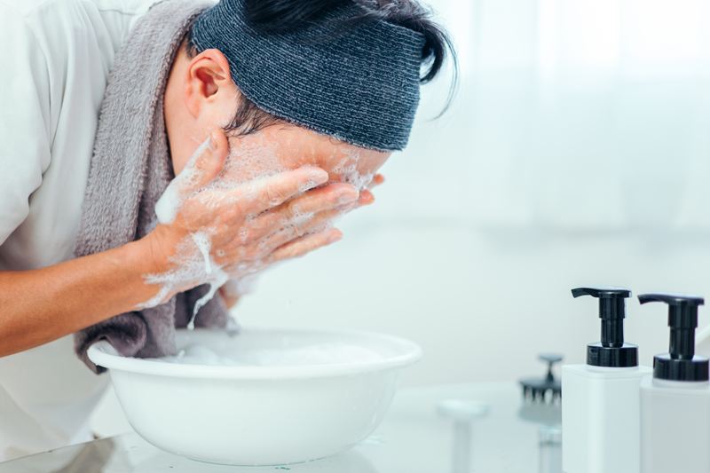 首にタオルをかけ、指先で泡を転がすように優しく洗顔する男性のアップ。